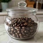 A pile of freshly roasted whole coffee beans in a glass jar on a kitchen counter.